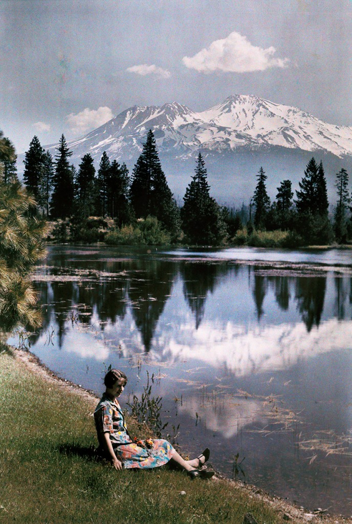 A girl sits by a lake with snow-capped mountains in the background, California, 1929